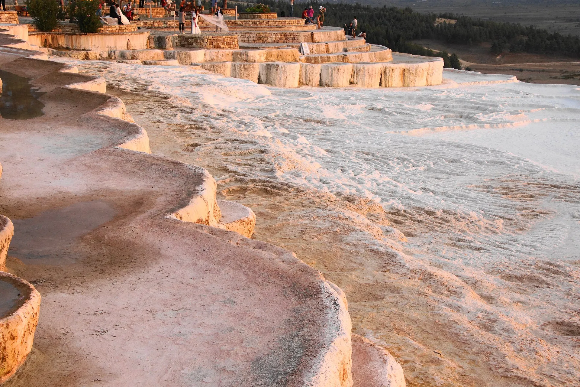 Visitors walking barefoot on Pamukkale's white travertine terraces with the ruins of Hierapolis on the hill above
